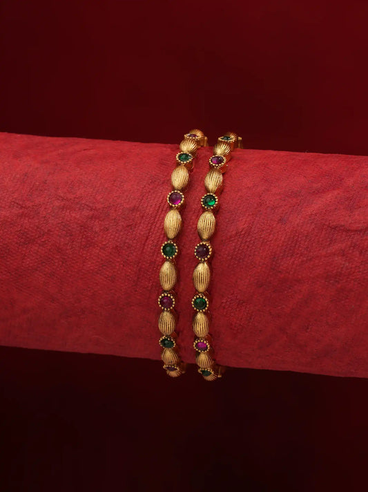 A pair of gold-plated bangles decorated with alternating oval textured beads and round green and pinkish-red gemstone accents, displayed on a textured red fabric roll against a dark red background.