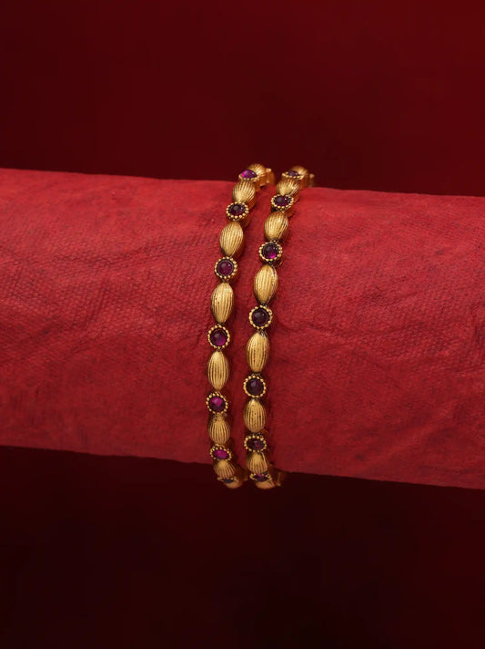 A pair of gold-plated traditional Indian bangles decorated with textured oval beads and small dark red gemstone accents, displayed on a textured red fabric stand.