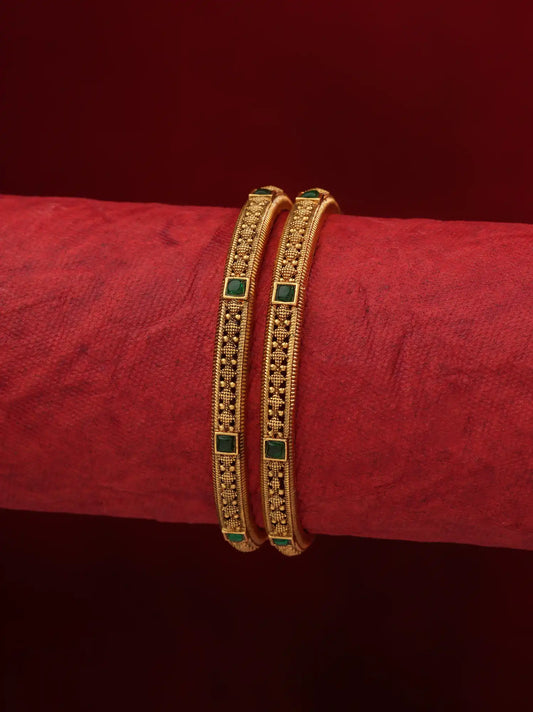 Two gold bangles with green stone accents, displayed on a textured red cylindrical stand against a dark red background.