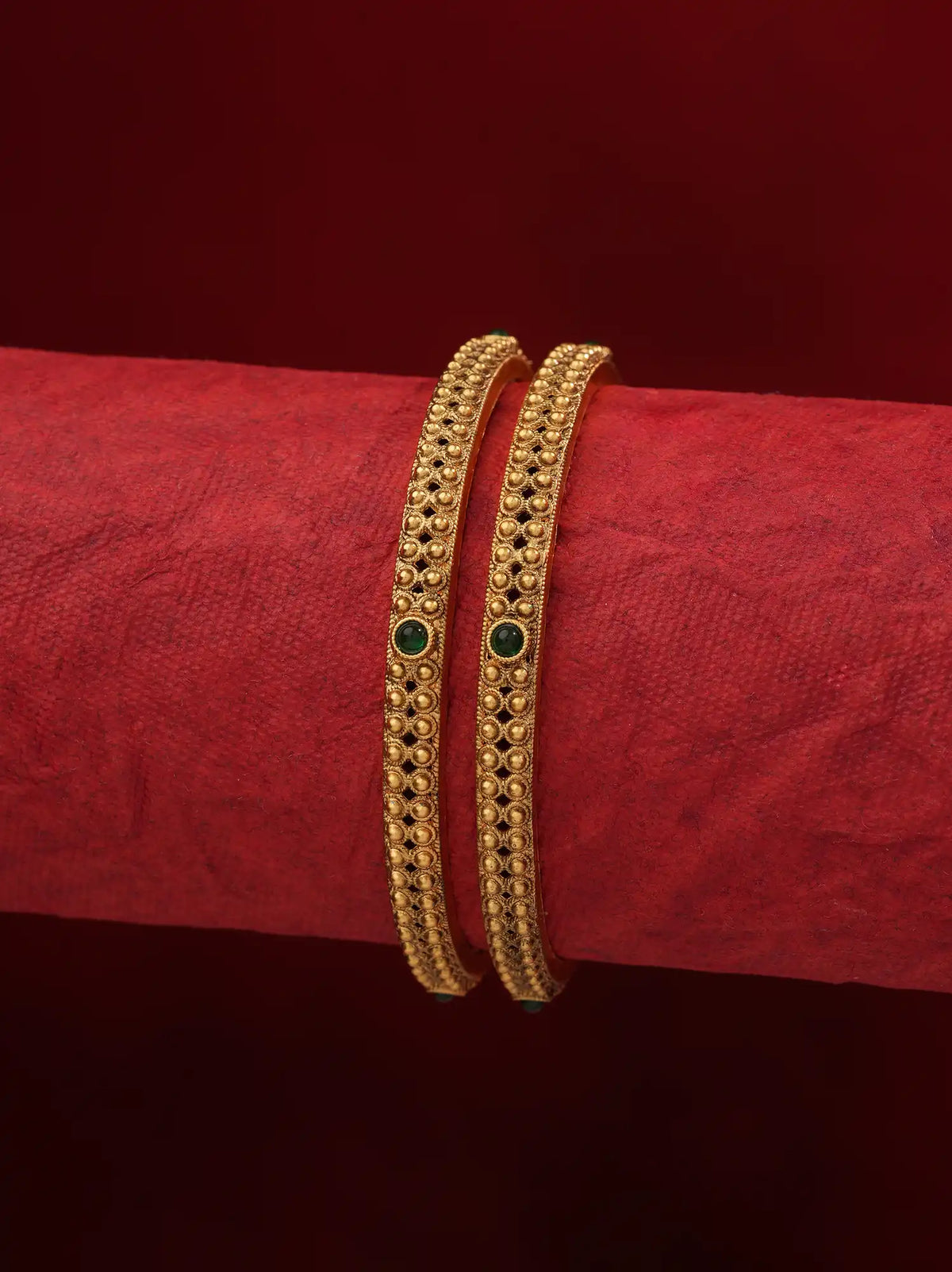 Two gold bangles with intricate filigree patterns and small green stone accents, displayed on a textured red cylindrical stand against a dark red background.