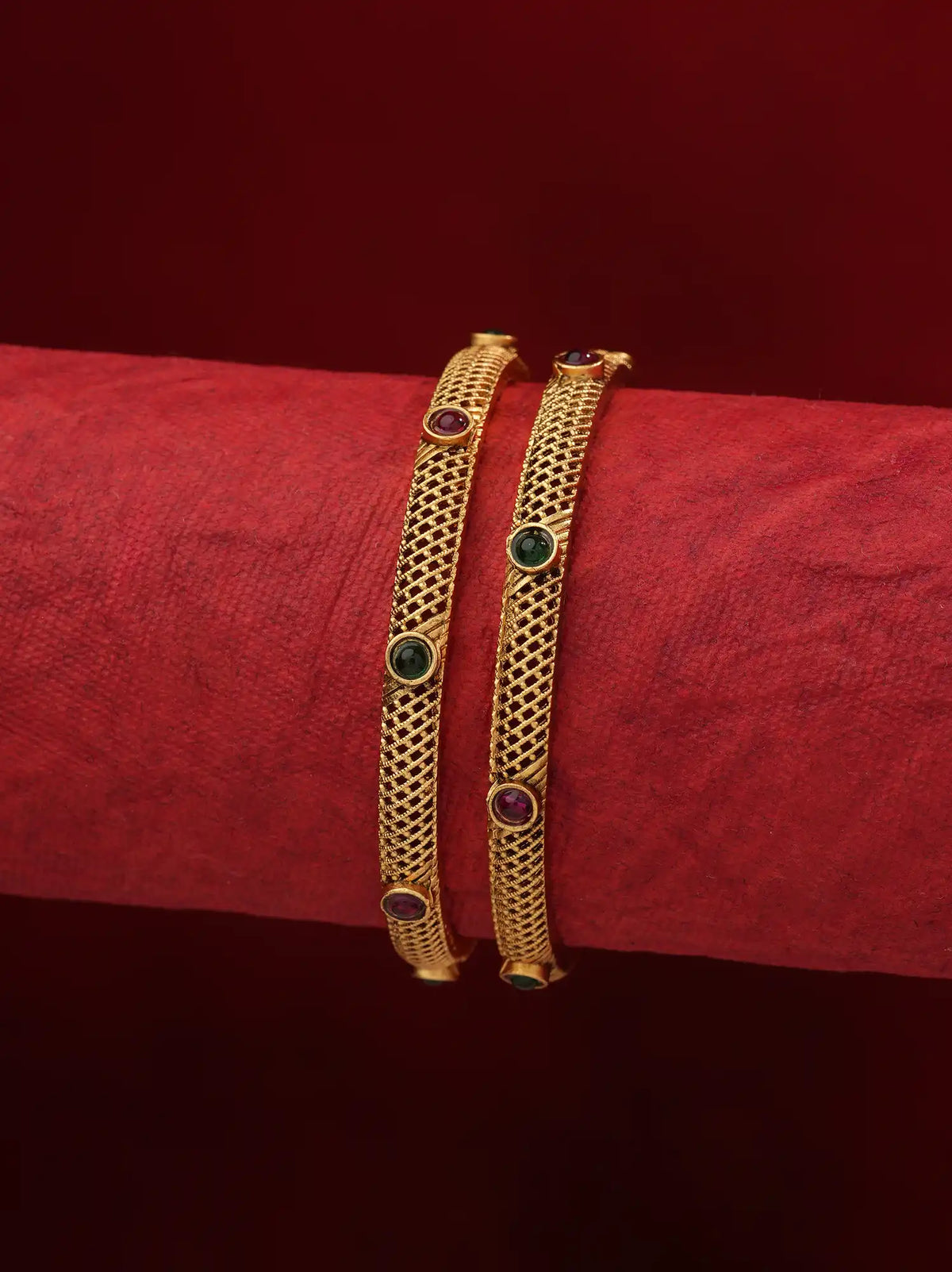 Two gold bangles with circular green and red stone accents, displayed on a textured red cylindrical stand against a dark red background.