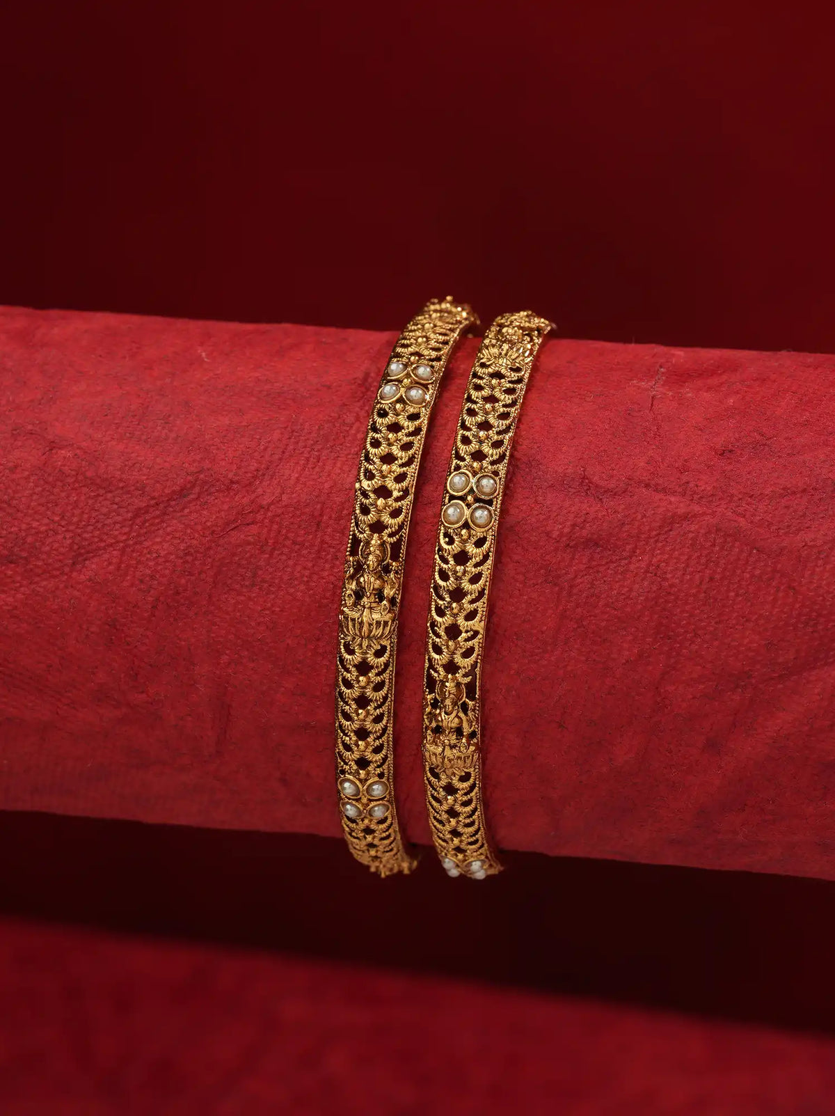 Two gold bangles with intricate filigree and small white stone accents, displayed on a textured red cylindrical stand against a matching red background.