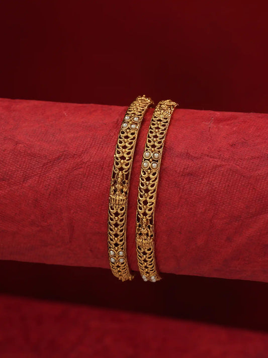 Two gold bangles with intricate filigree and small white stone accents, displayed on a textured red cylindrical stand against a matching red background.