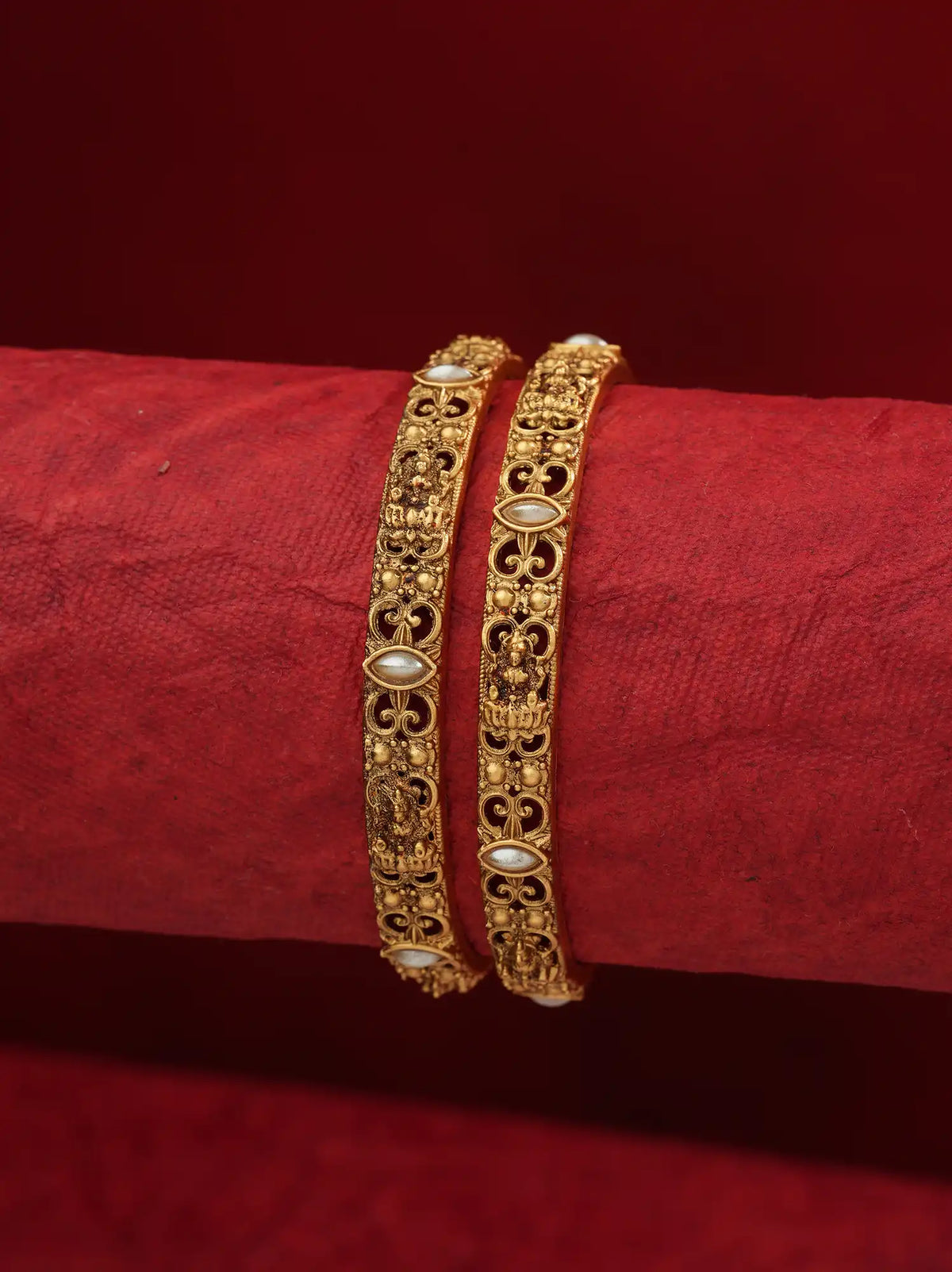 Two ornate gold bangles with white pearl-like accents, displayed on a red textured cylindrical stand against a dark red background.