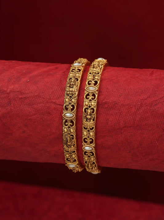 Two ornate gold bangles with white pearl-like accents, displayed on a red textured cylindrical stand against a dark red background.