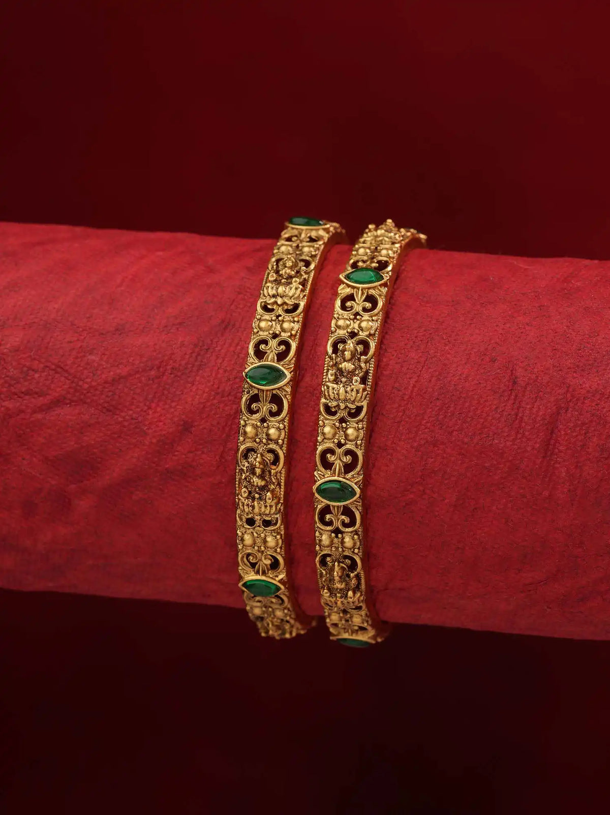 Two ornate gold bangles with green stone accents, displayed on a red textured surface against a dark red background.