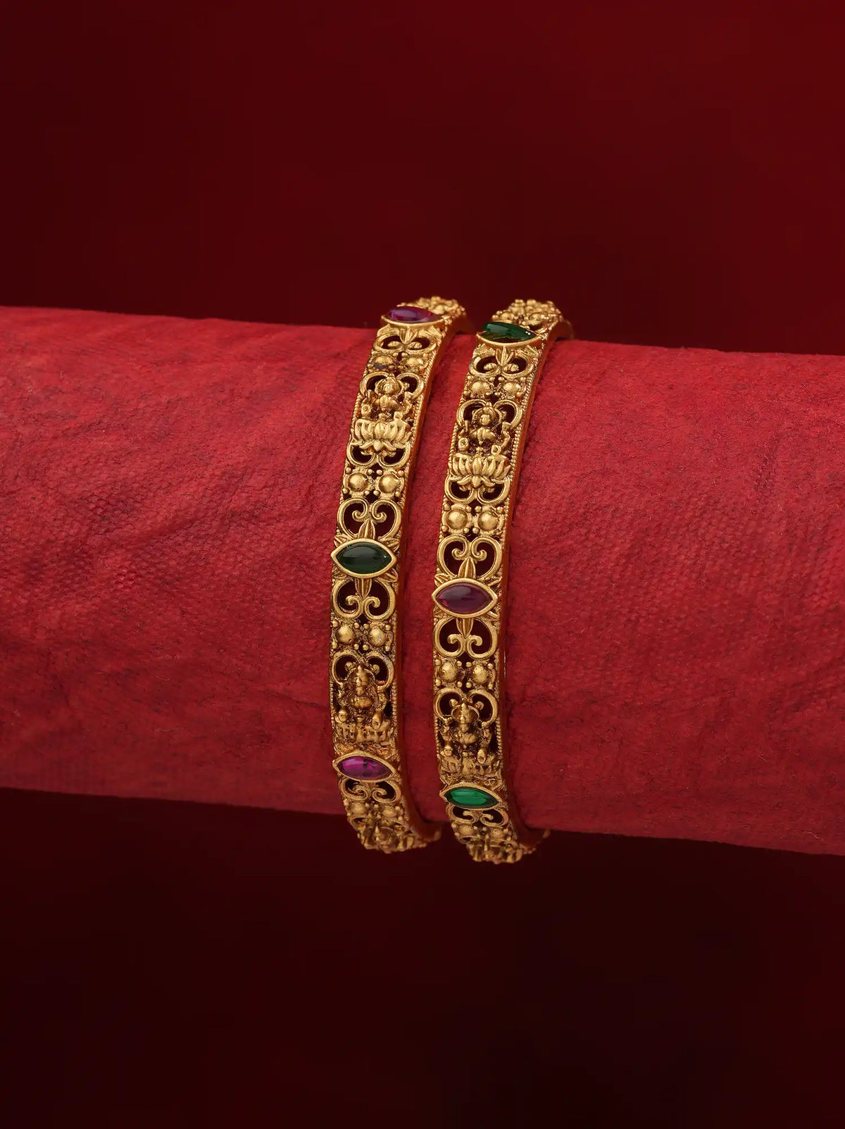 Two ornate gold bangles with intricate filigree work and small oval green and purple stone accents, displayed on a red fabric-wrapped wrist against a dark red background.