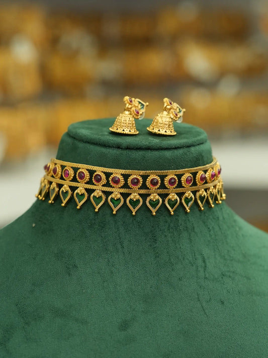 Gold-toned traditional Indian jewelry set featuring a choker necklace and matching bell-shaped earrings, embellished with red and green stones, displayed on a dark green velvet jewelry bust.