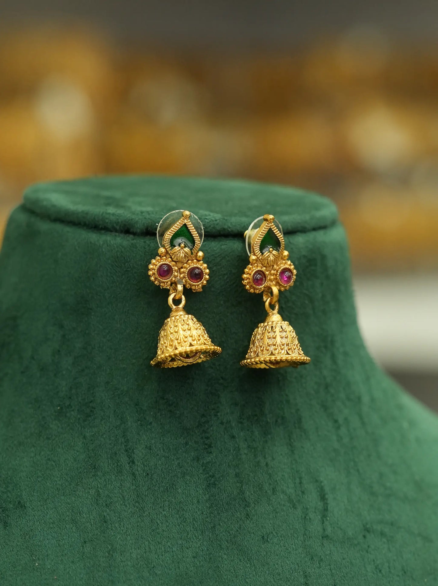 A pair of gold-toned traditional Indian jhumka earrings with red and green stone accents, displayed on a dark green velvet jewelry bust against a blurred golden background.