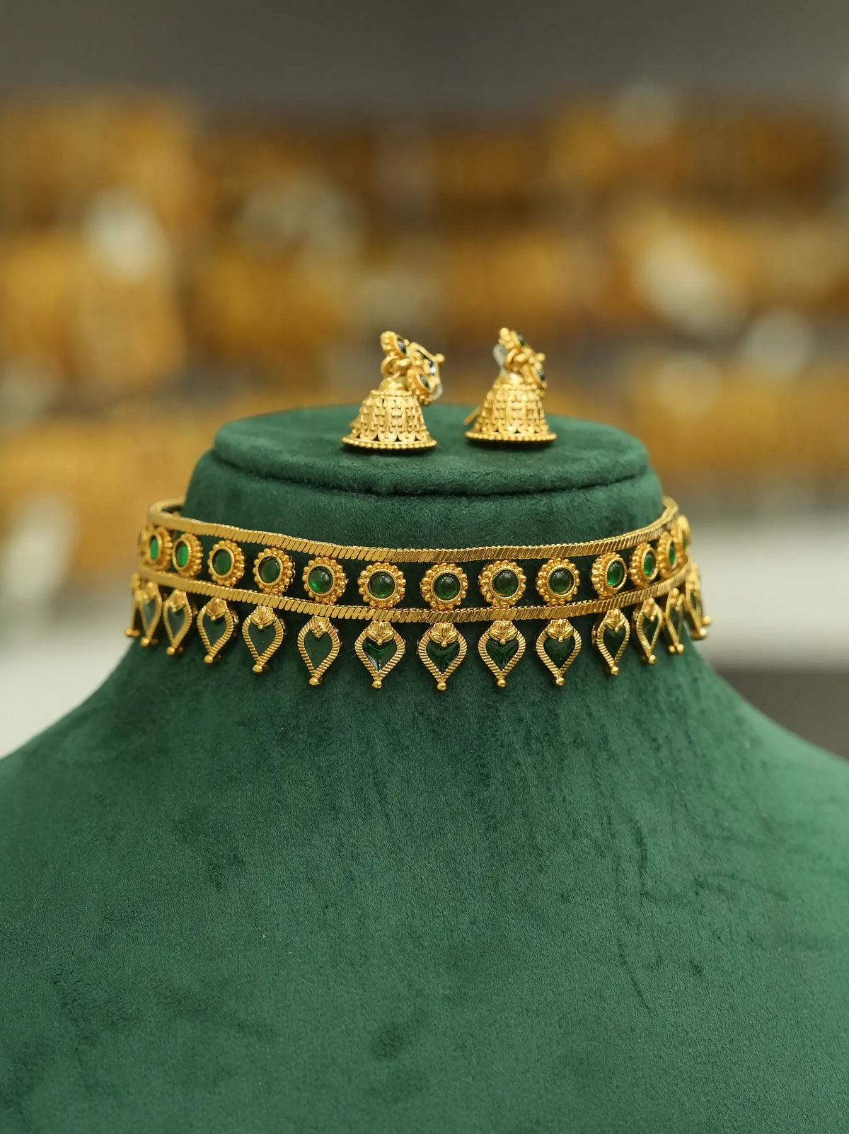 Gold-toned traditional jewelry set displayed on a dark green velvet jewelry bust, featuring a choker necklace with green stone accents and dangling heart motifs, plus matching bell-shaped earrings.