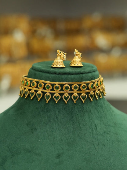 Gold-toned traditional jewelry set displayed on a dark green velvet jewelry bust, featuring a choker necklace with green stone accents and dangling heart motifs, plus matching bell-shaped earrings.