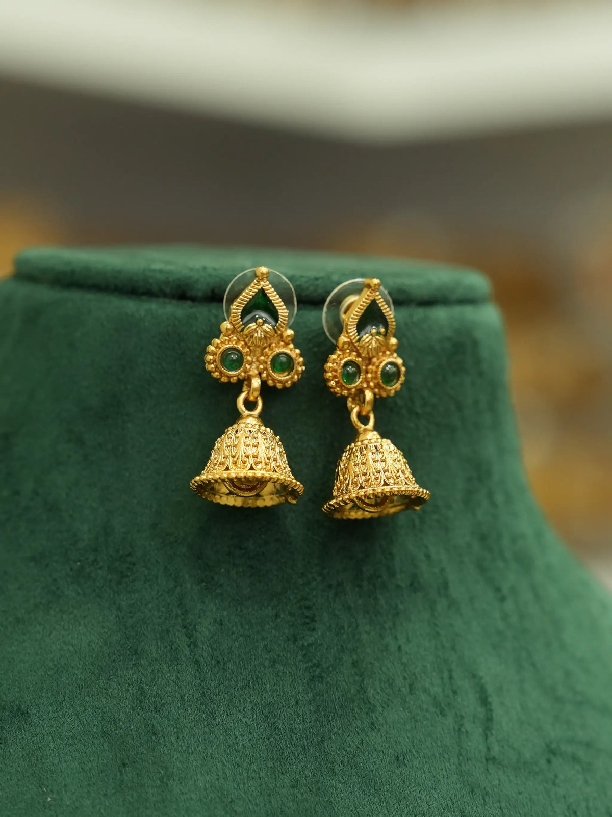 A pair of gold-toned traditional jhumka earrings with green stone accents, displayed on a dark green velvet jewelry bust.
