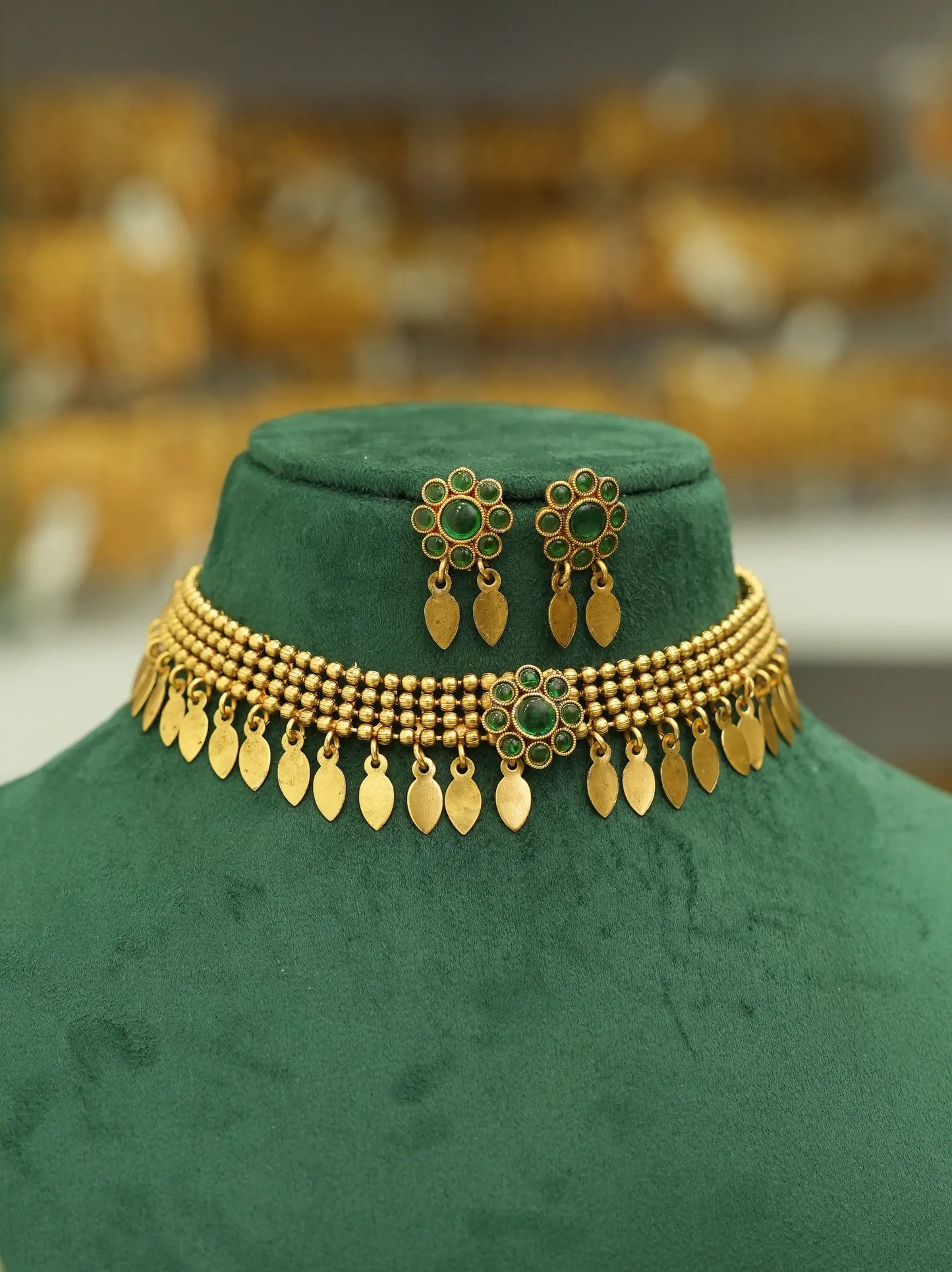 Gold-toned traditional jewelry set displayed on a dark green velvet mannequin bust, including a beaded necklace with dangling leaf-shaped charms, matching earrings, and a small pendant, against a softly blurred warm-toned background.