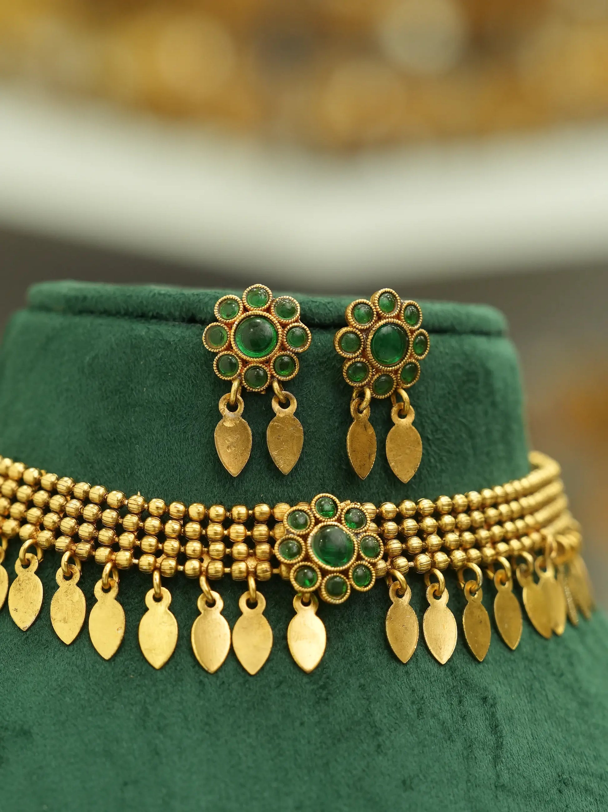 Gold-toned traditional jewelry set featuring a beaded necklace and matching earrings with green stone accents, displayed on a dark green velvet jewelry stand.