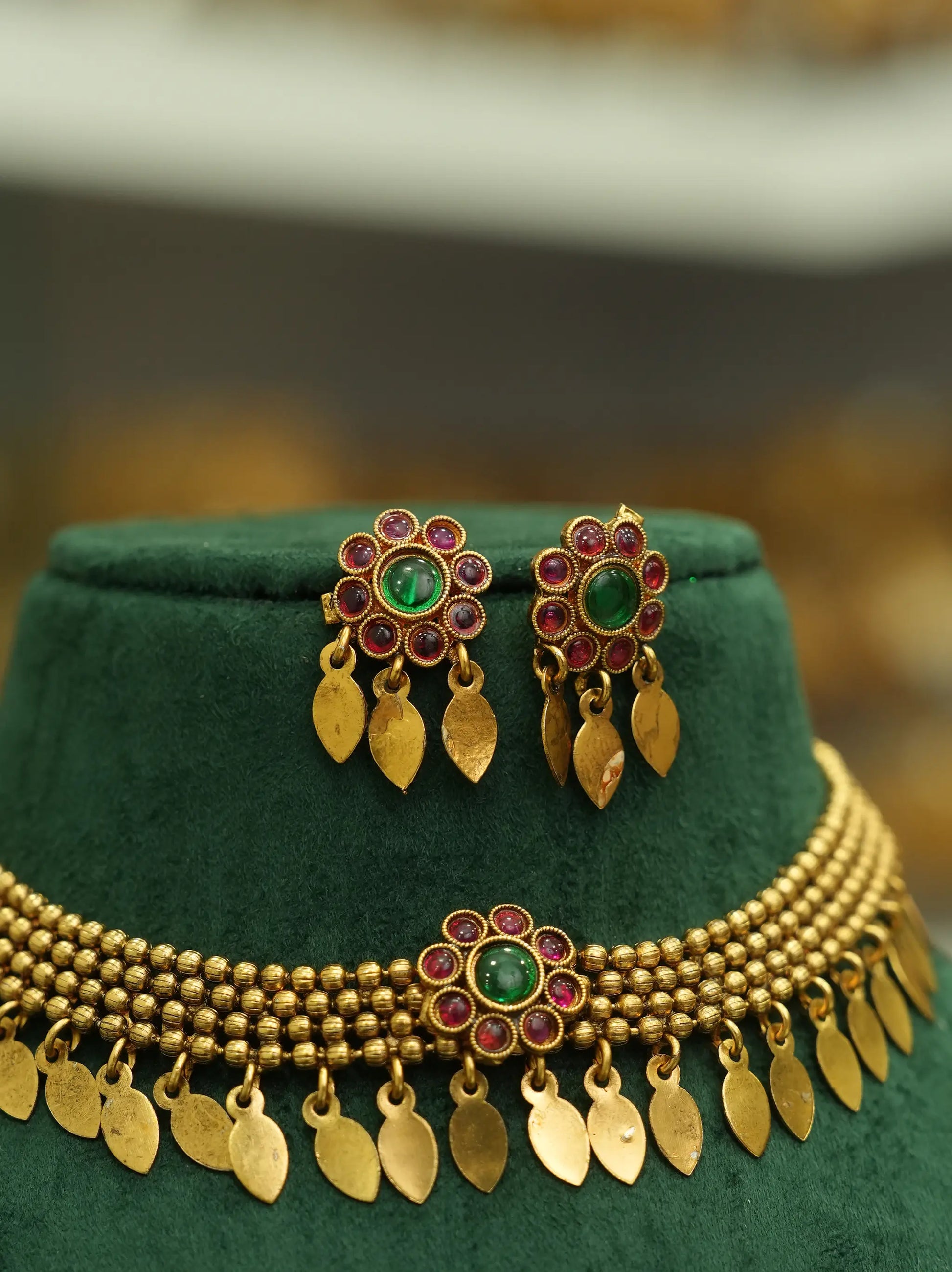 Gold-toned traditional jewelry set including a necklace and matching earrings displayed on a dark green velvet stand. The pieces feature gold leaf-shaped dangling charms, beaded strands, and central floral motifs with green oval stones surrounded by small red stones.