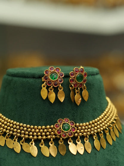 Gold-toned traditional jewelry set including a necklace and matching earrings displayed on a dark green velvet stand. The pieces feature gold leaf-shaped dangling charms, beaded strands, and central floral motifs with green oval stones surrounded by small red stones.