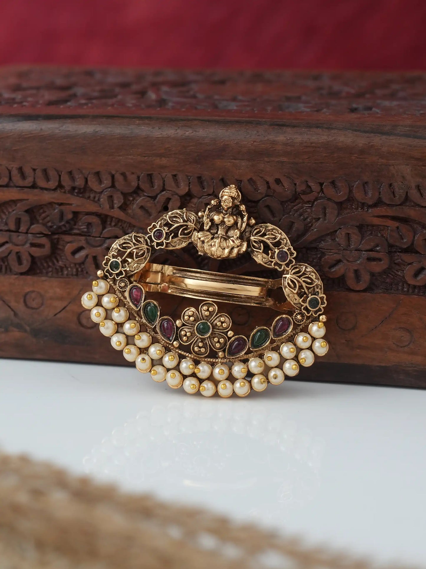 An ornate gold-toned traditional Indian brooch featuring a deity motif, a curved row of white faux pearls, and small green and red enamel accents, displayed on a carved wooden box.
