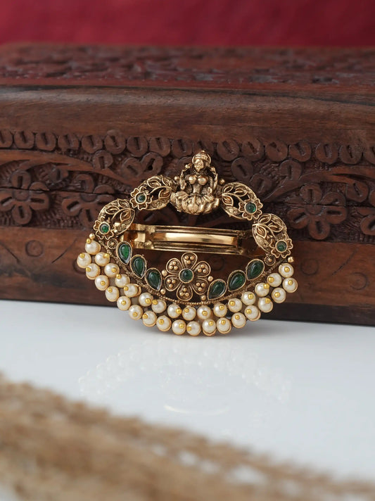 An ornate gold-toned traditional Indian brooch with green stone accents and a row of white faux pearls, displayed on a white surface in front of a carved dark wood box.