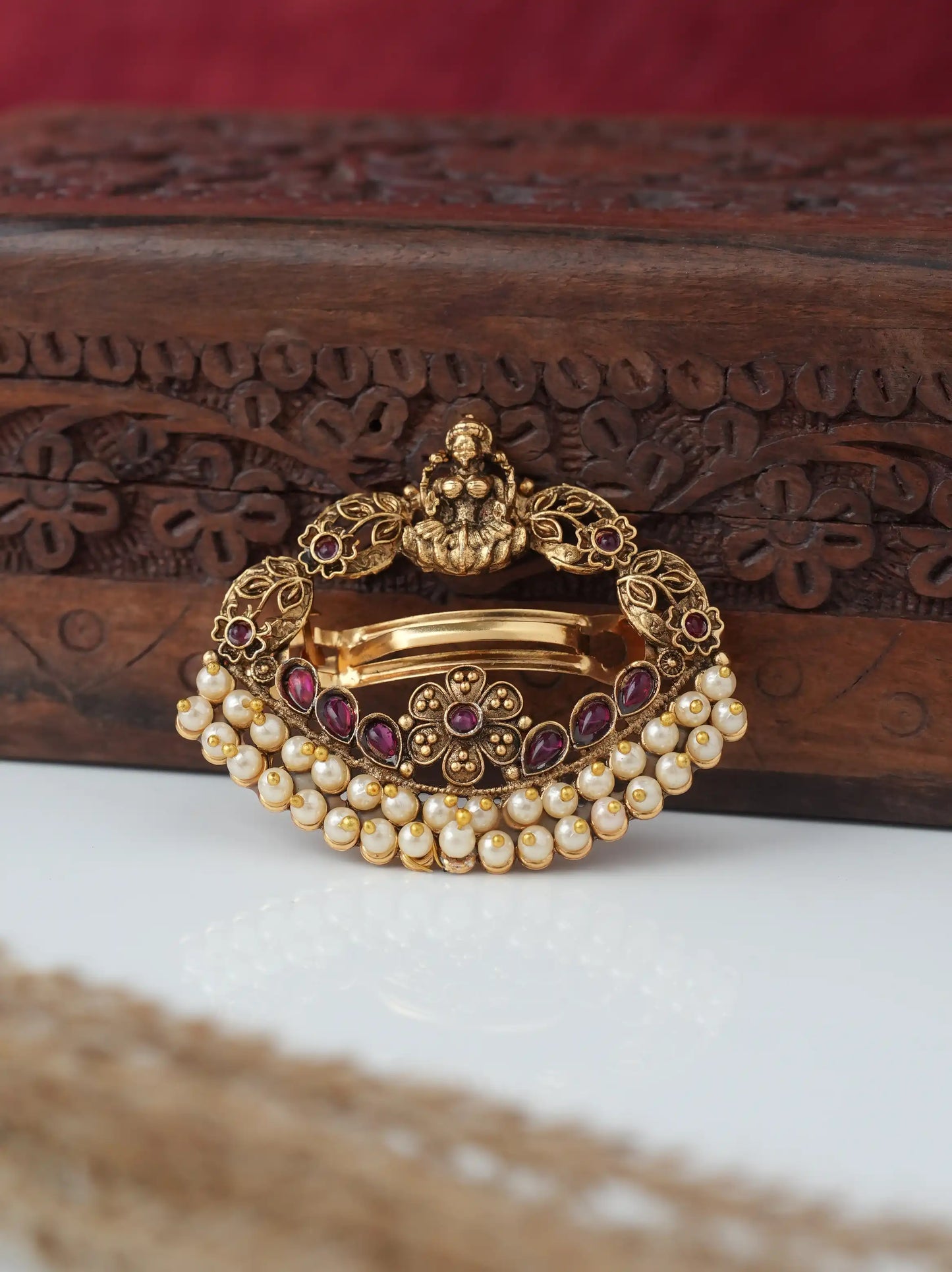 An ornate gold-toned traditional Indian brooch decorated with white pearls, red gemstones, and a central gold motif, displayed on a carved wooden box.