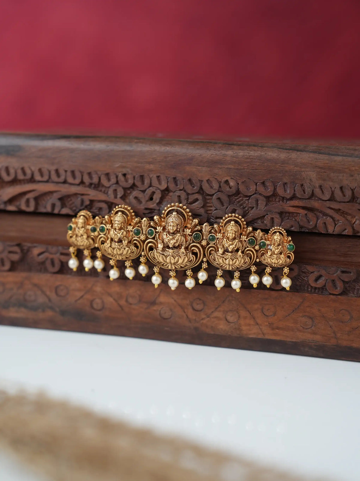 An ornate gold-toned traditional Indian hair ornament (jhumka-style) with three central deity motifs, small green stone accents, and dangling white pearl beads, displayed on a carved dark wood surface against a red and white background.