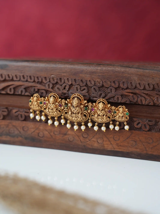 A gold-toned traditional Indian jewelry piece featuring three Ganesha motifs, small green and red stone accents, and dangling pearl beads, displayed on a carved dark wood surface against a red background.
