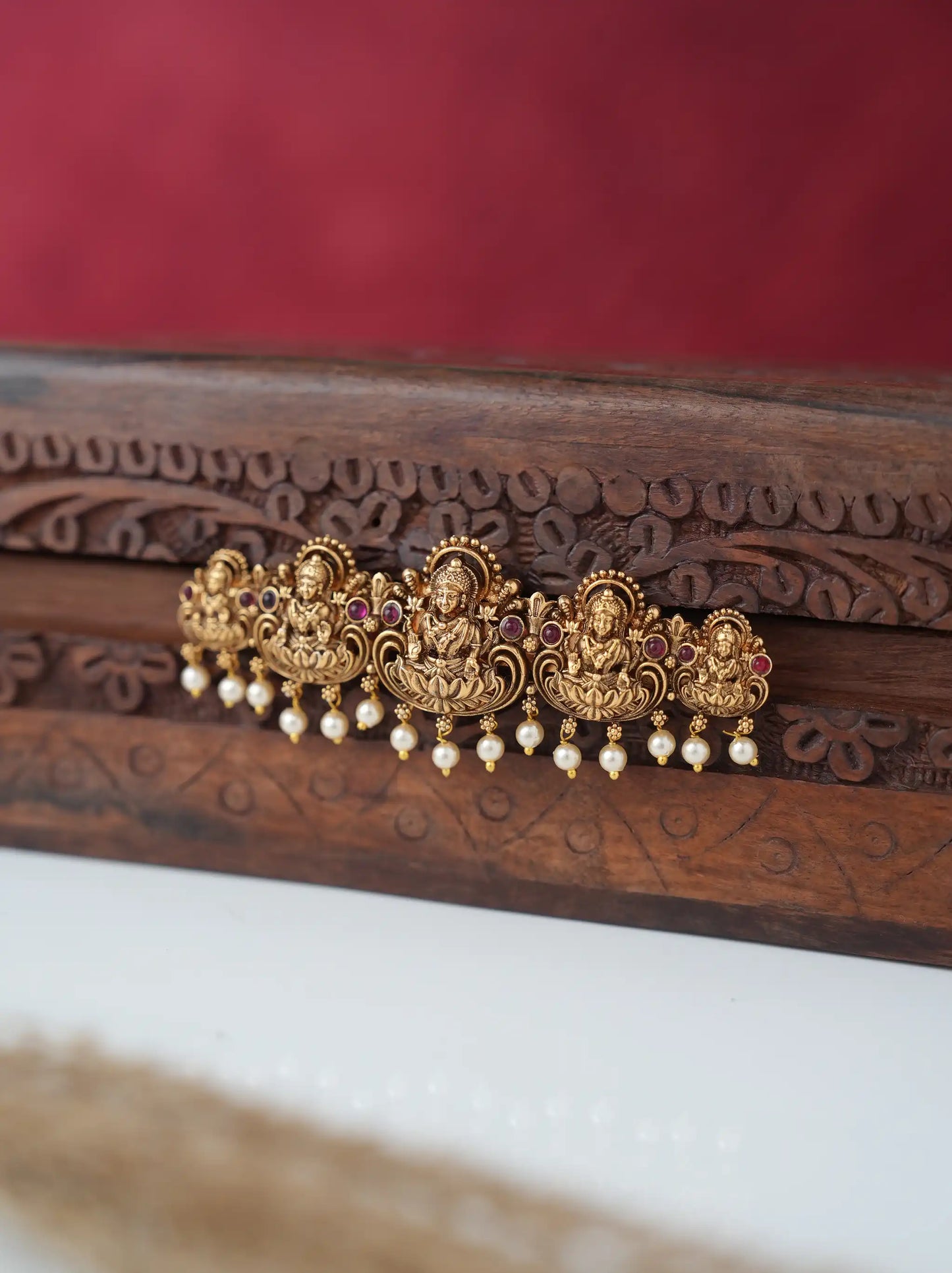 An ornate gold-toned traditional Indian hair ornament (jhumka) with red stone accents and dangling white pearl beads, displayed on a carved dark wood surface against a red background.