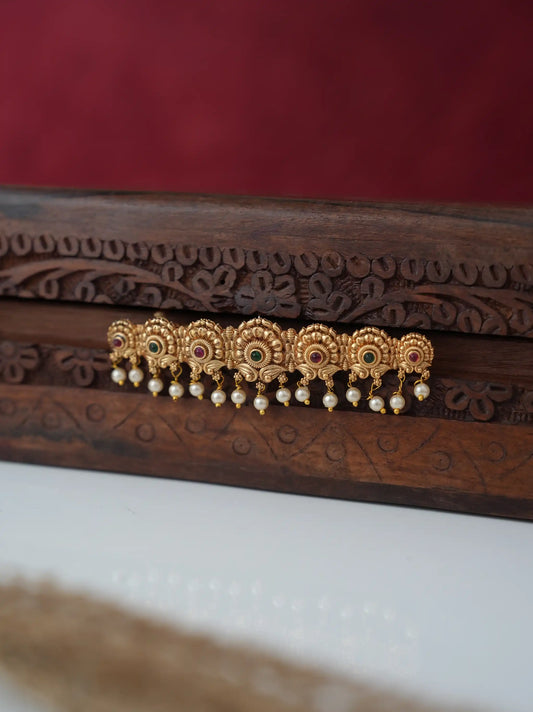 A gold-toned traditional Indian hair ornament (jhumka) with small colorful stone accents and dangling white pearls, displayed on a carved dark wooden box against a dark red background.