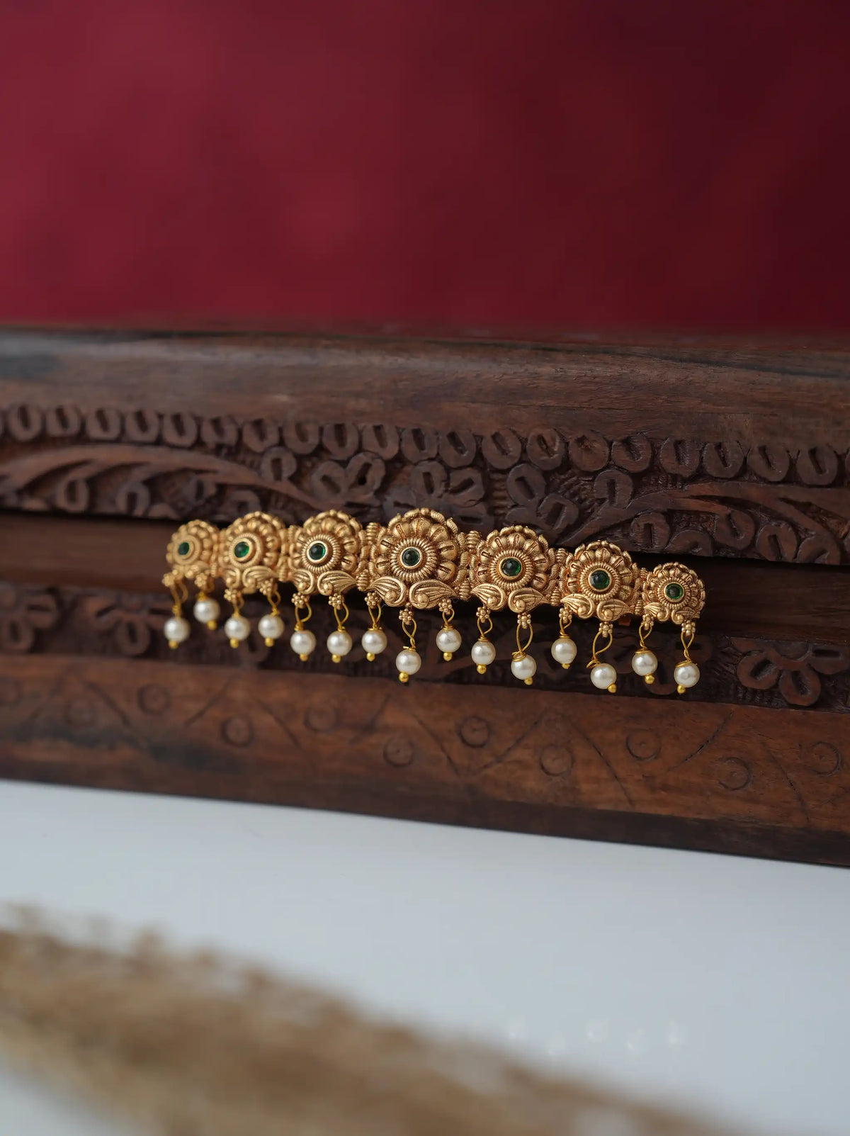 A gold-toned traditional Indian hair ornament (jhumka) with green stone accents and dangling white pearl beads, displayed on a carved dark wood surface against a dark red background.
