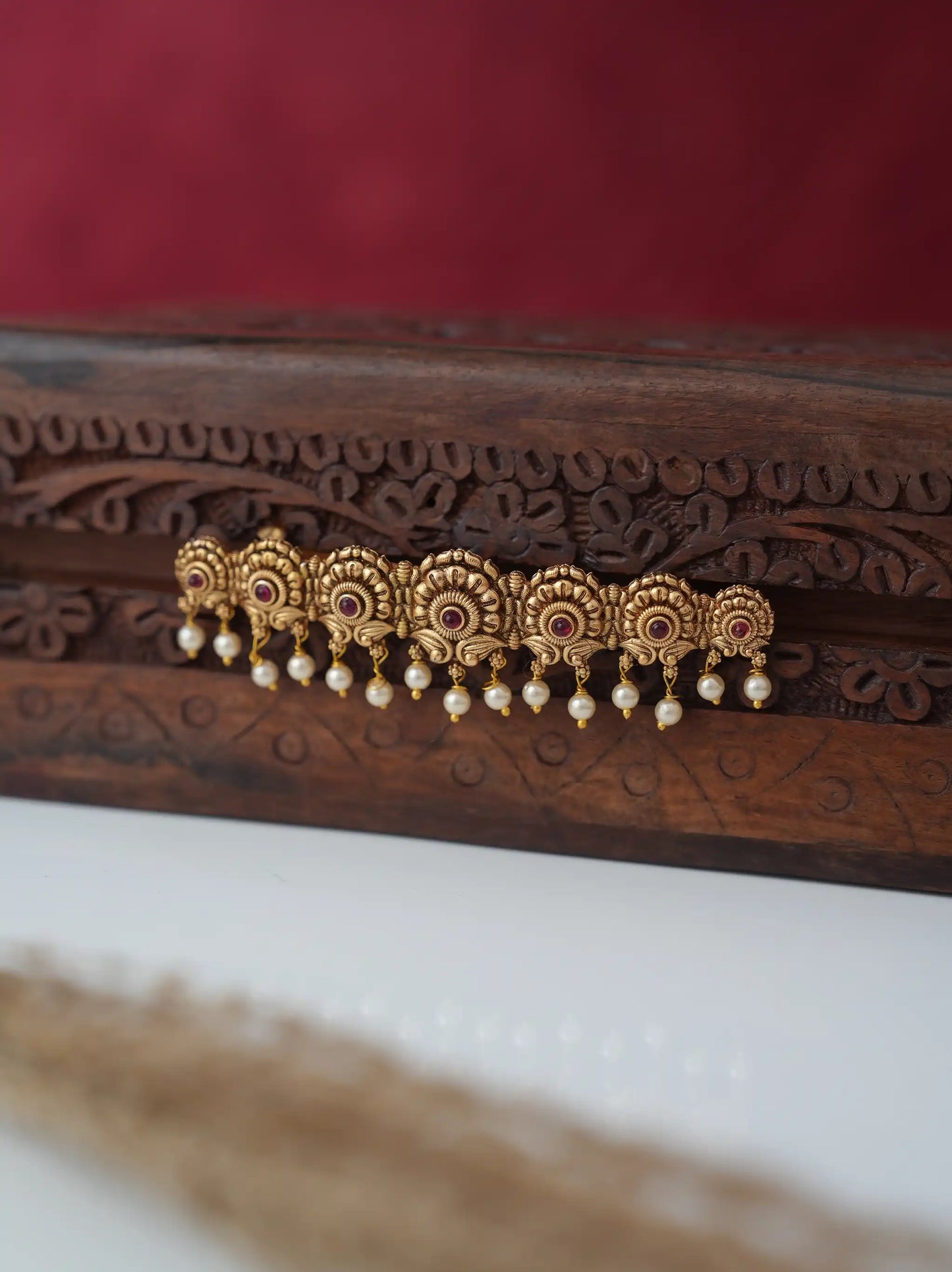 A close-up of ornate gold-toned traditional earrings with small red stone accents and dangling white pearl drops, displayed on a carved dark wooden surface against a deep red background.