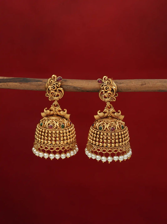 Gold-toned peacock jhumka earrings with pearl drops, close-up angled view showing filigree dome and pearl beading, on a wooden stand against a maroon background