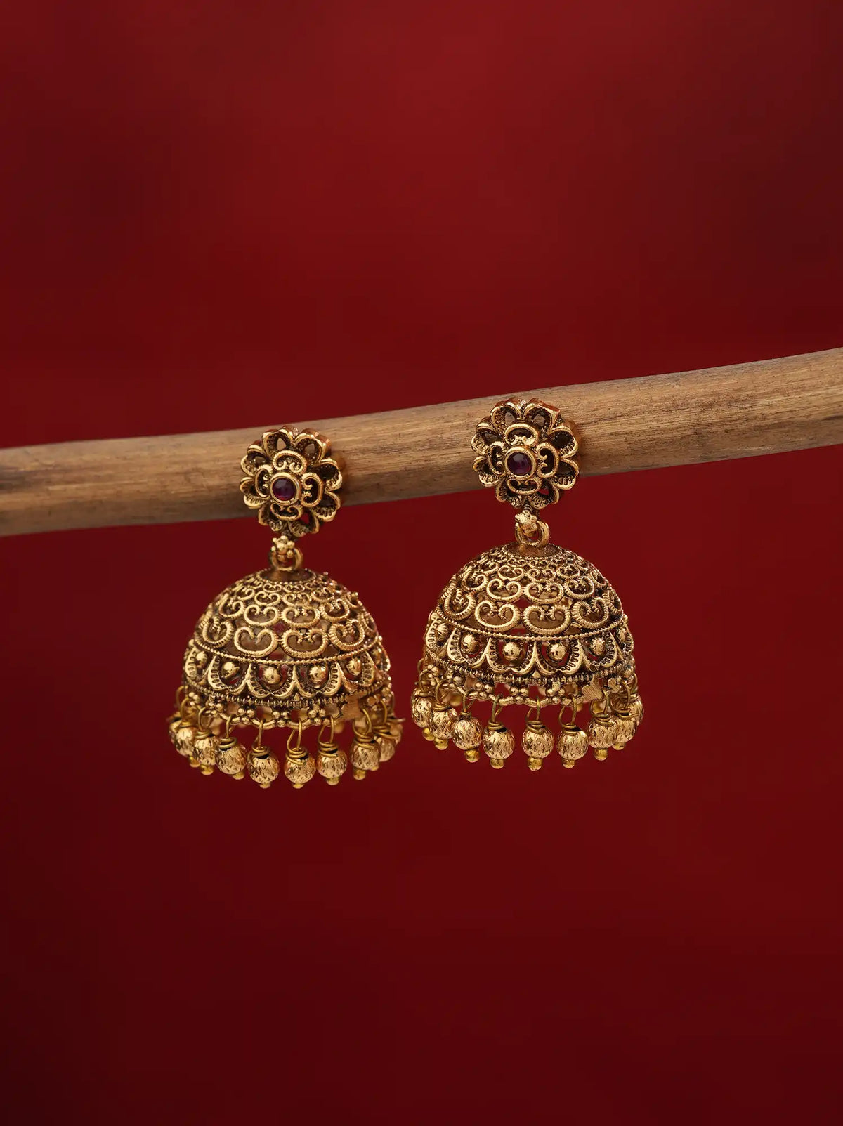 A pair of ornate gold-toned jhumka earrings with intricate filigree detailing and small dark stone accents, hanging from a wooden branch against a solid deep red background.