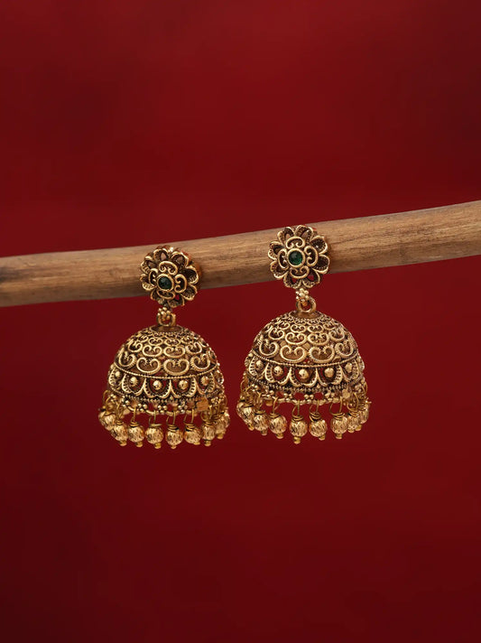 A pair of ornate gold-toned jhumka earrings with small green stone accents and dangling bead chains, hanging from a wooden branch against a solid dark red background.