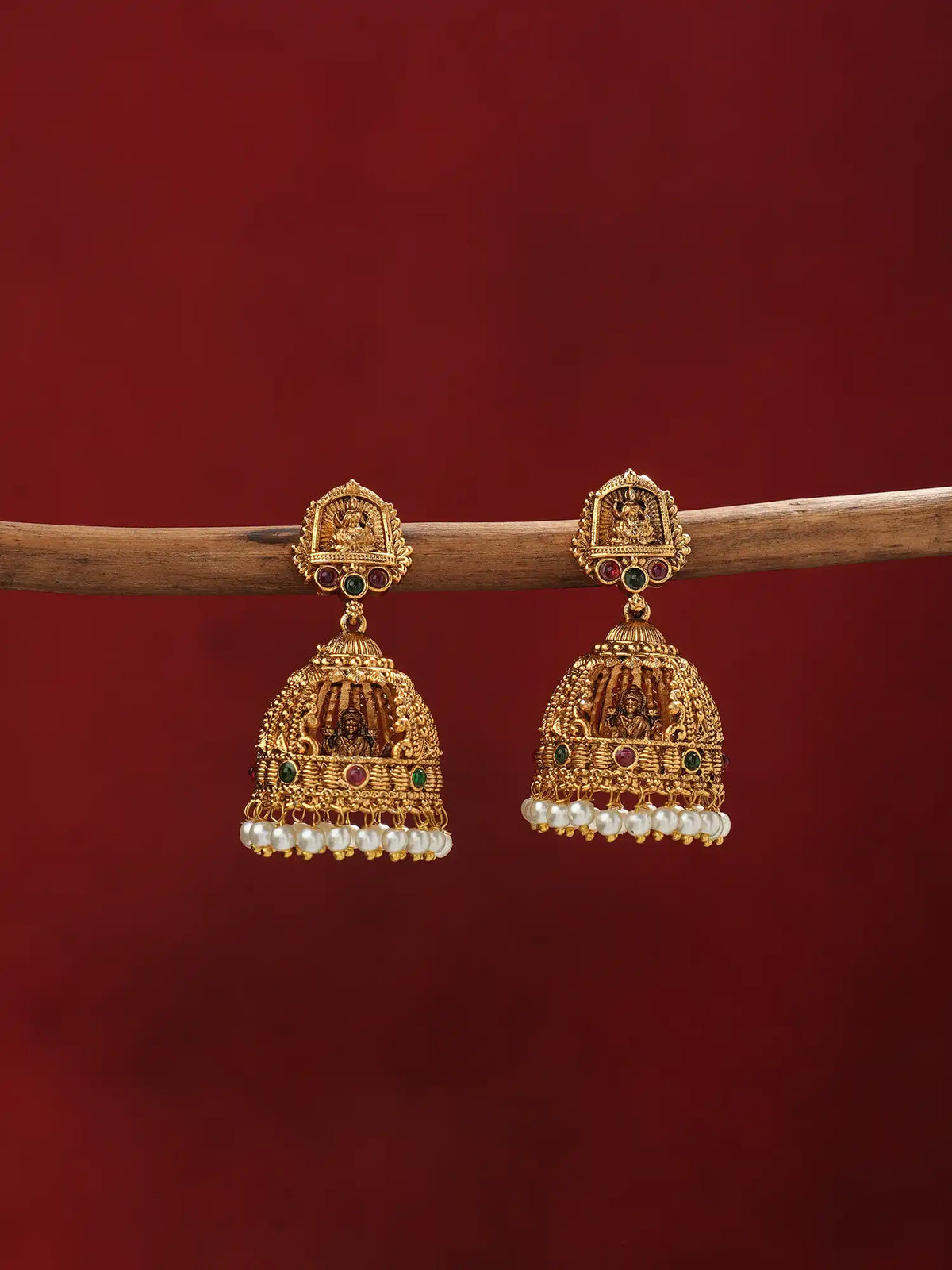 A pair of ornate gold-toned Indian jhumka earrings with purple bead fringe, hanging from a wooden branch against a dark red background.