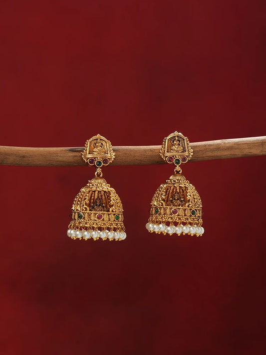 A pair of ornate gold-toned Indian jhumka earrings with purple bead fringe, hanging from a wooden branch against a dark red background.