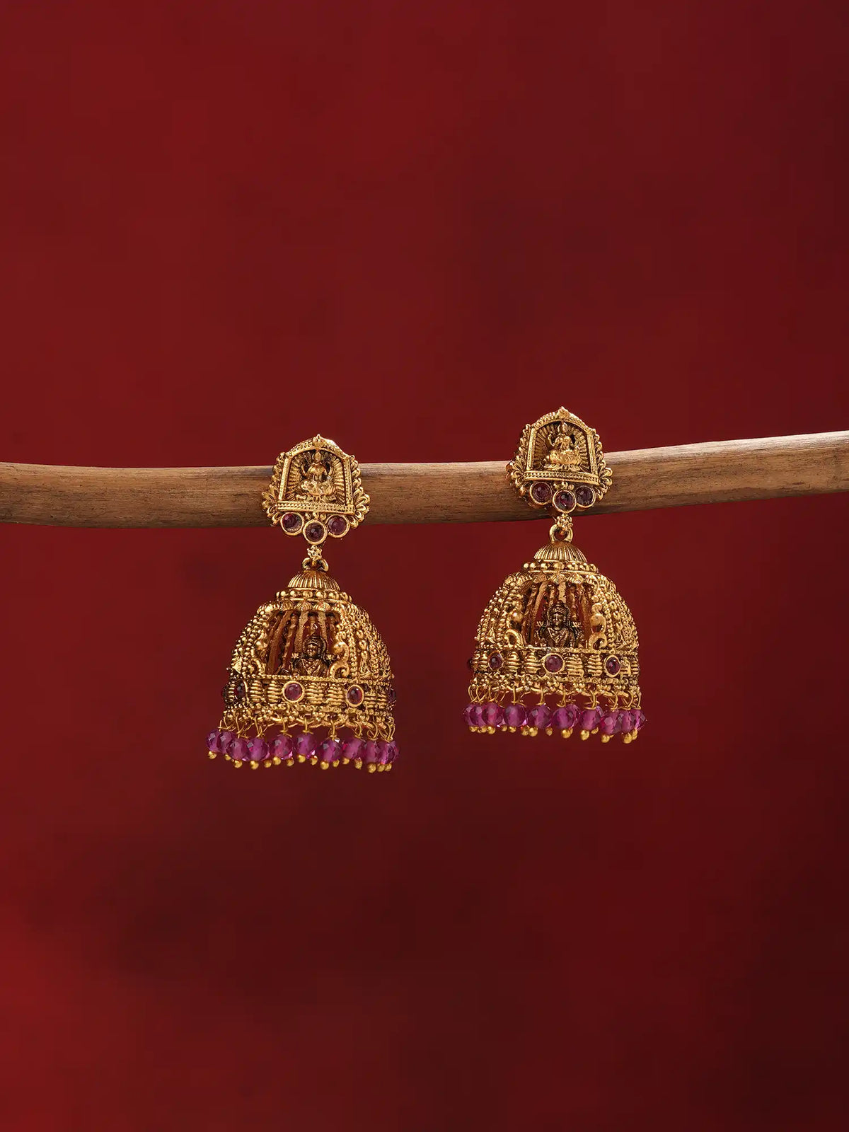 A pair of ornate gold-toned Indian jhumka earrings with purple bead fringe, hanging from a wooden branch against a solid dark red background.