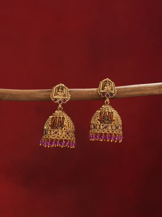 A pair of ornate gold-toned Indian jhumka earrings with purple bead fringe, hanging from a wooden branch against a solid dark red background.