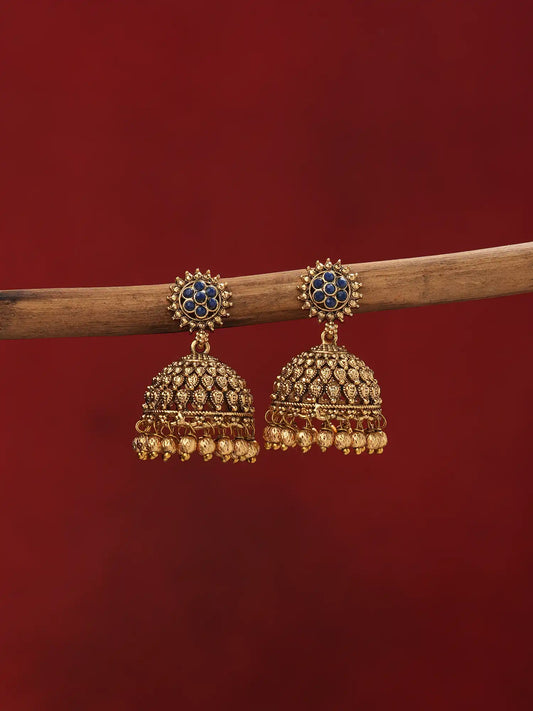A pair of traditional Indian gold jhumka earrings with blue stone accents and dangling gold bells, hanging from a wooden branch against a solid dark red background.