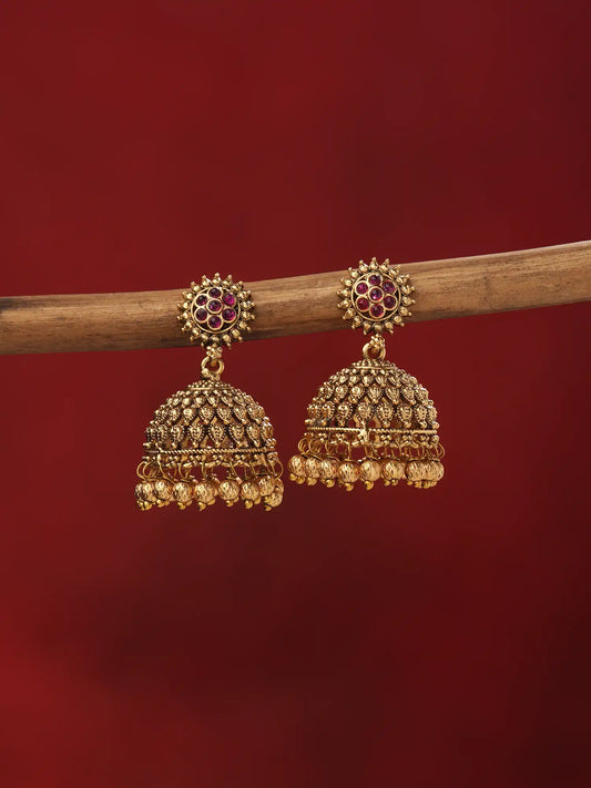 A pair of ornate gold-toned jhumka earrings with red and dark stone detailing, hanging from a wooden branch against a solid deep red background.