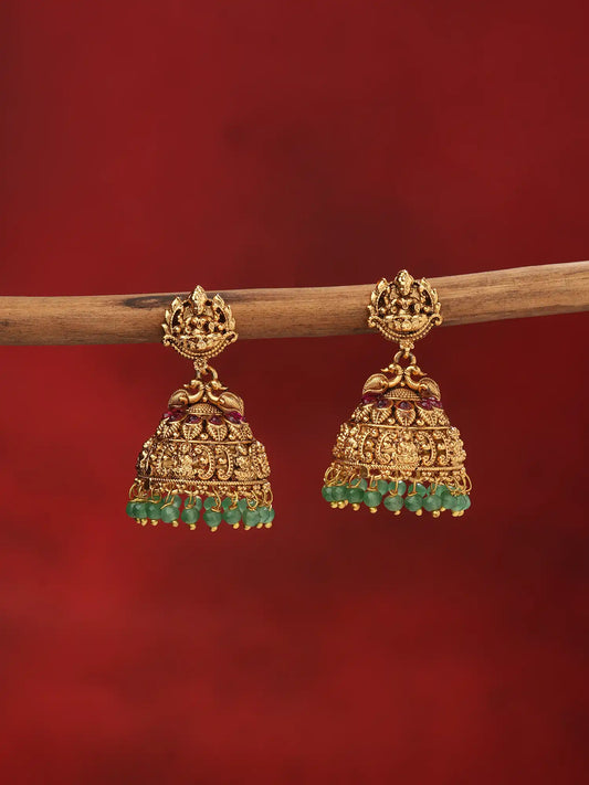 A pair of traditional Indian gold jhumka earrings with green stone accents and small purple stone details, hanging from a wooden branch against a solid dark red background.