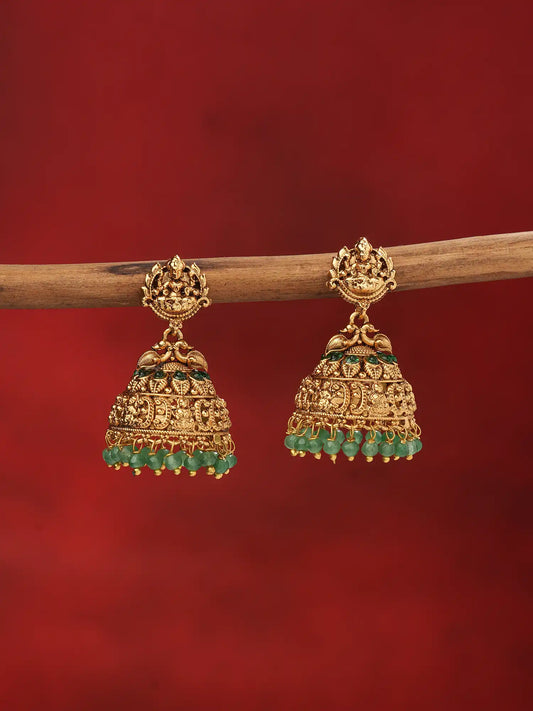 A pair of traditional Indian gold jhumka earrings with green stone accents, hanging from a wooden branch against a solid dark red background.