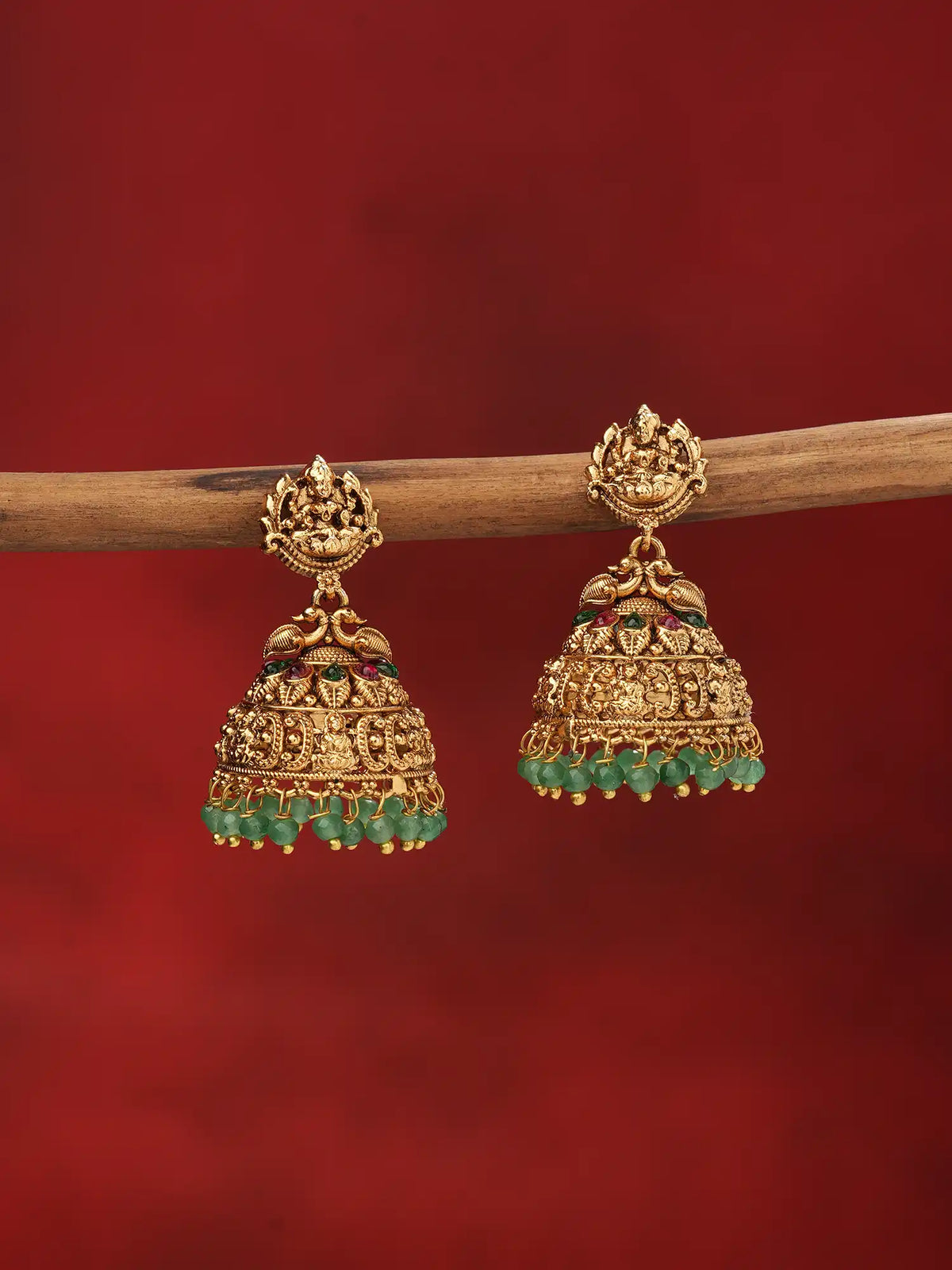 A pair of traditional Indian gold jhumka earrings with green bead fringe and small red stone accents, hanging from a wooden branch against a solid dark red background.
