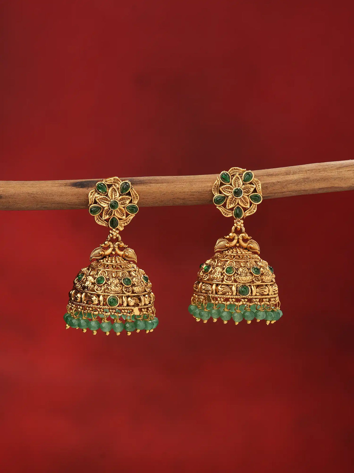 A pair of gold-toned traditional Indian jhumka earrings embellished with green stones and hanging green bead tassels, displayed on a wooden branch against a solid dark red background.