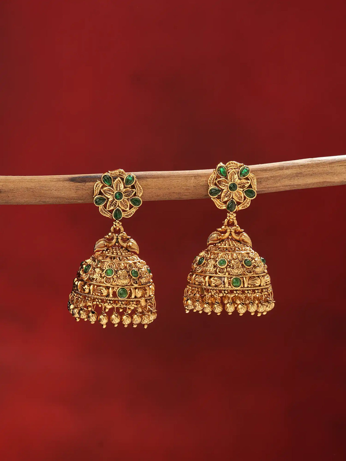 A pair of ornate gold jhumka earrings with green stone accents, hanging from a wooden branch against a solid dark red background.