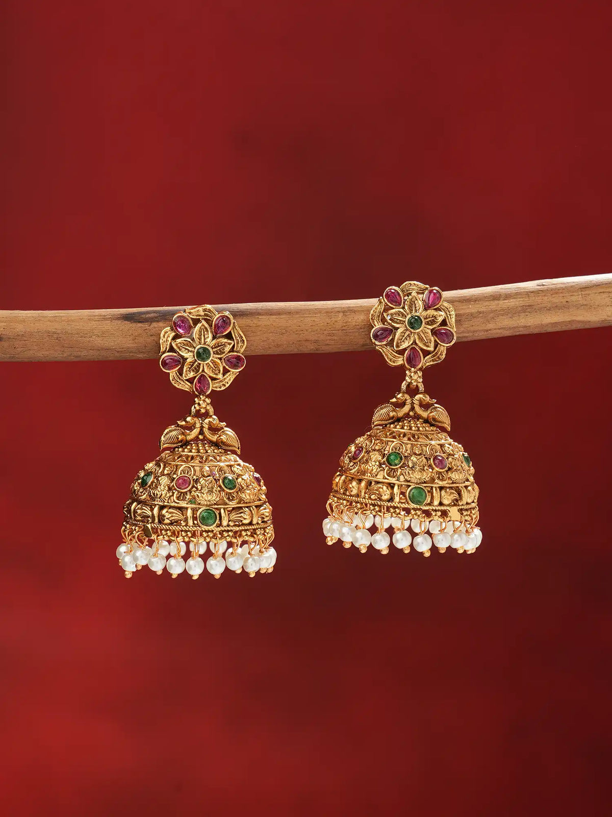 A pair of ornate gold-toned Indian jhumka earrings with red and green stones and white pearl fringe, hanging from a wooden branch against a dark red background.