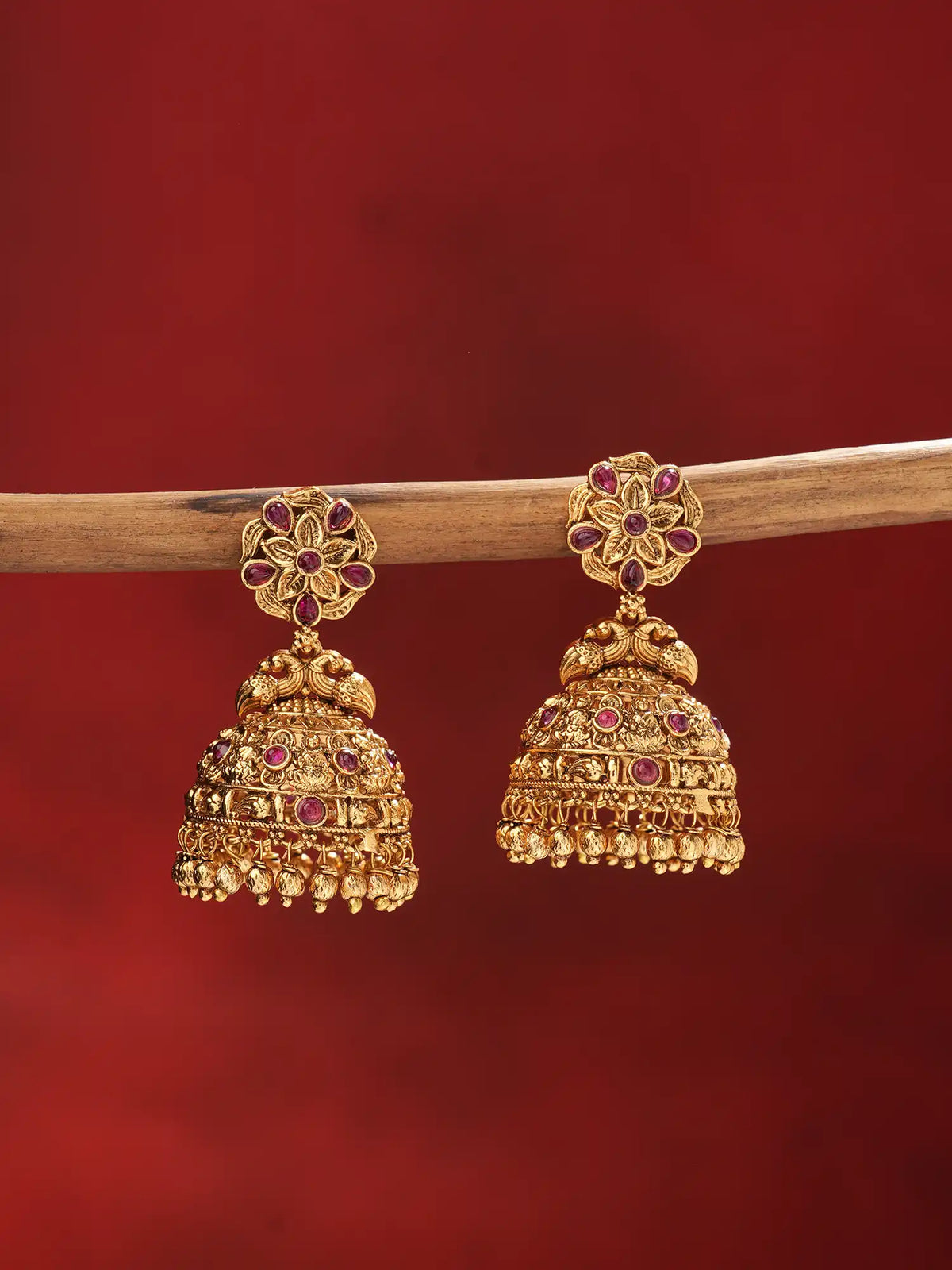 A pair of ornate gold-toned jhumka earrings with red gemstone accents, hanging from a wooden branch against a solid dark red background.