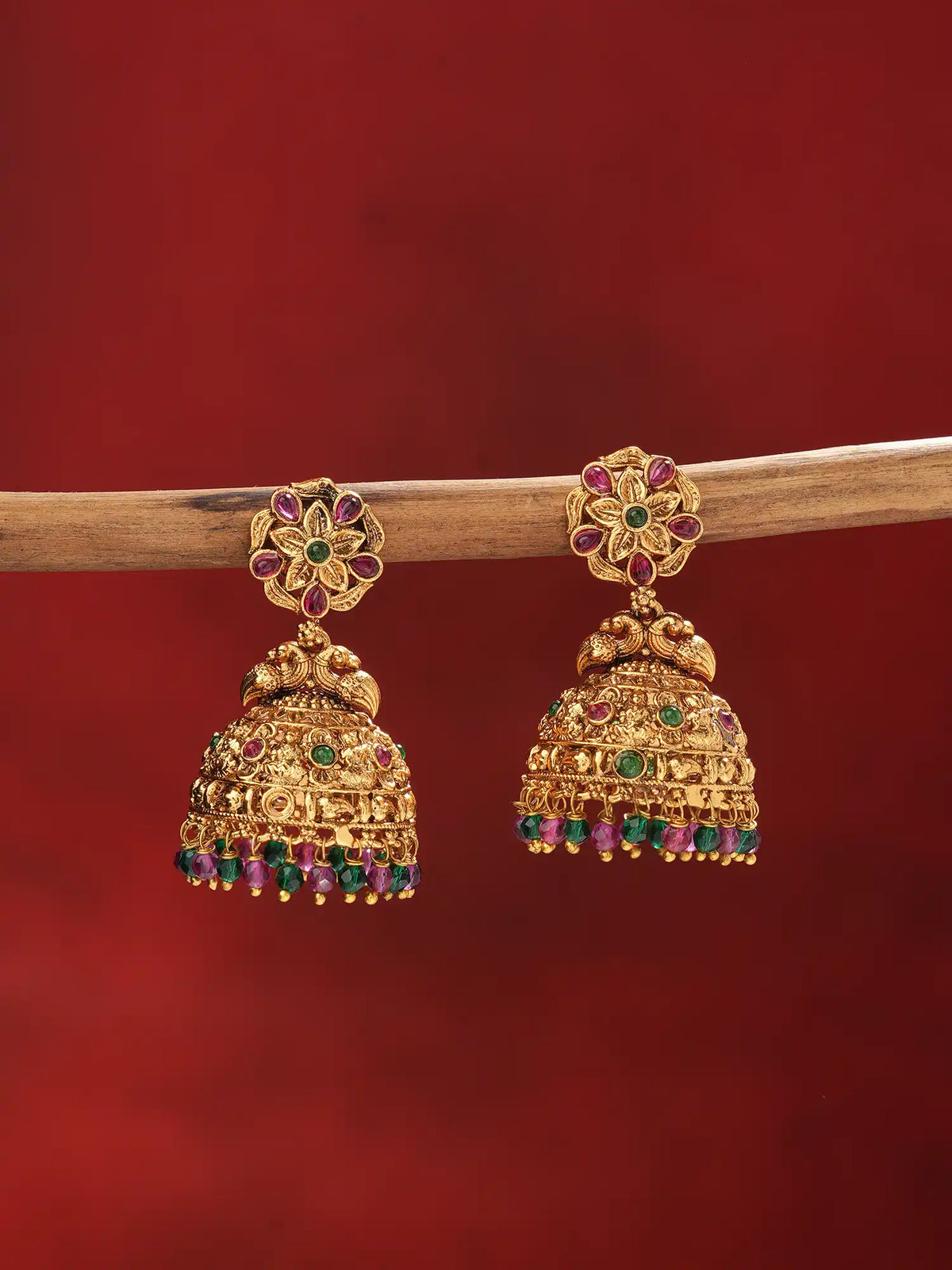 A pair of ornate gold jhumka earrings with red and green stone accents, hanging from a wooden branch against a solid dark red background.