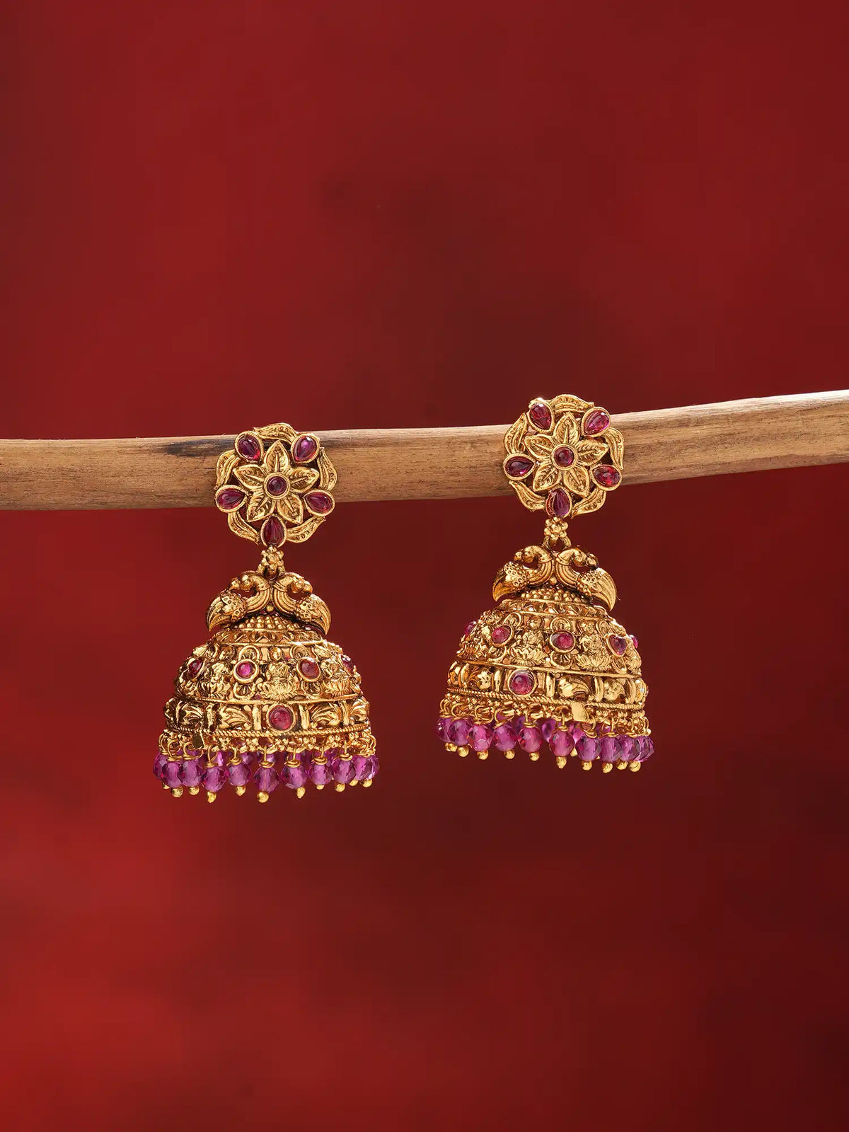 A pair of ornate gold-toned jhumka earrings with red gemstone accents, hanging from a wooden branch against a deep red background.