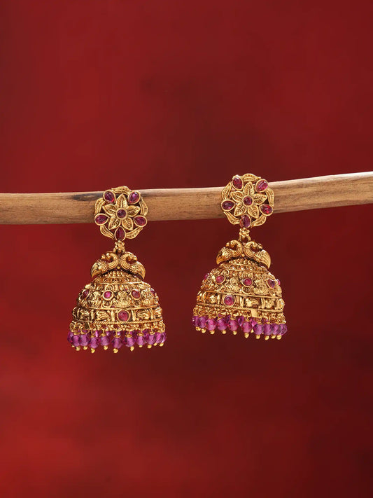 A pair of ornate gold-toned jhumka earrings with red gemstone accents, hanging from a wooden branch against a deep red background.