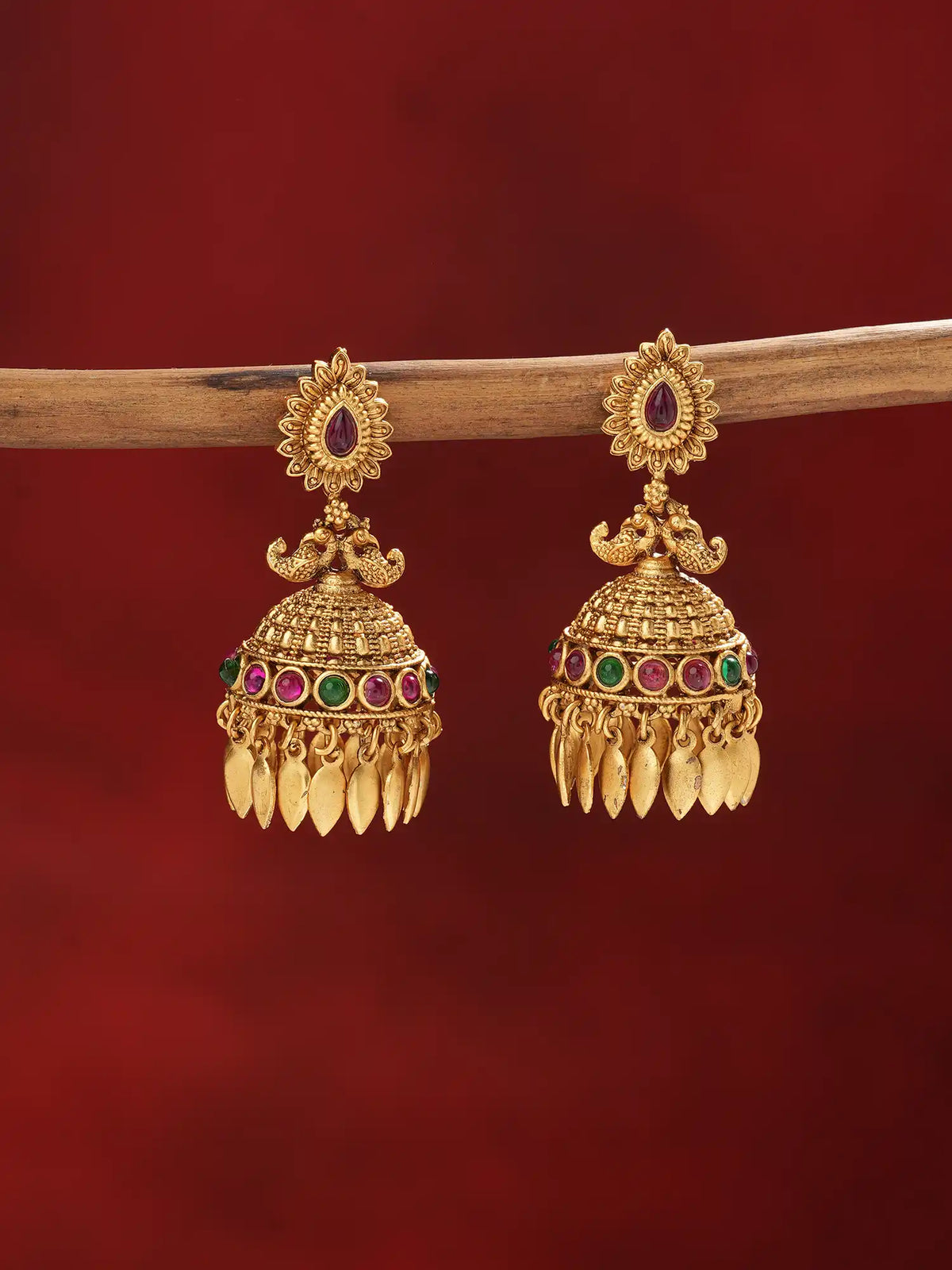 A pair of ornate gold-toned Indian jhumka earrings with colorful stone accents and dangling leaf-shaped charms, hanging from a wooden branch against a dark red background.