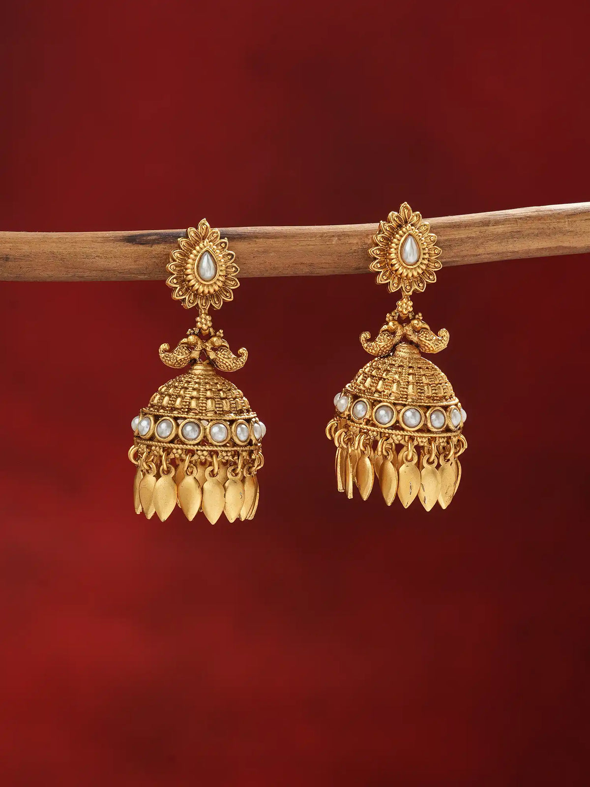 A pair of ornate gold-toned Indian jhumka earrings with white pearl-like accents, hanging from a wooden branch against a deep red background.
